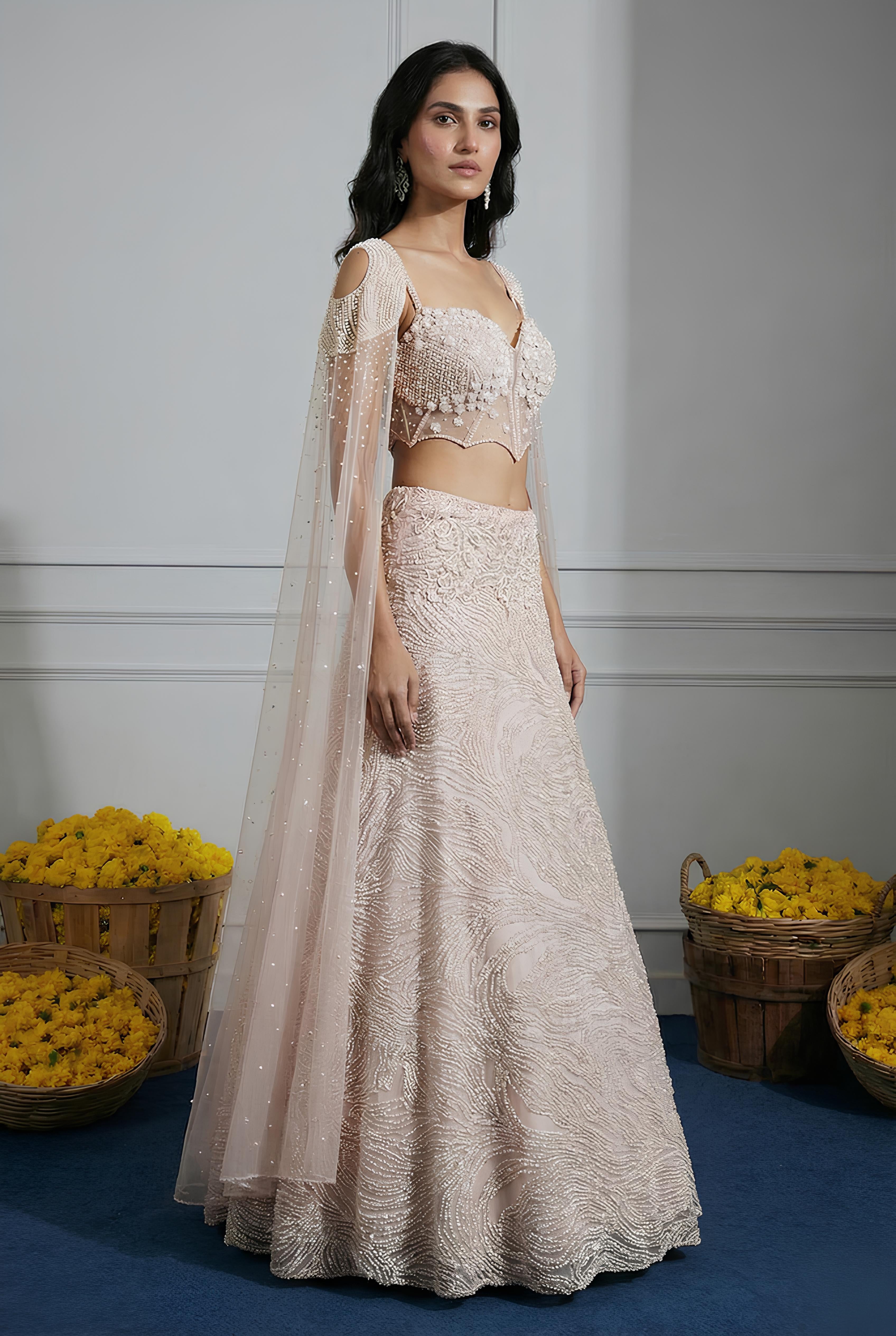 Woman in a beige embroidered lehenga with a matching dupatta standing against a white wall with yellow flowers in the background.
